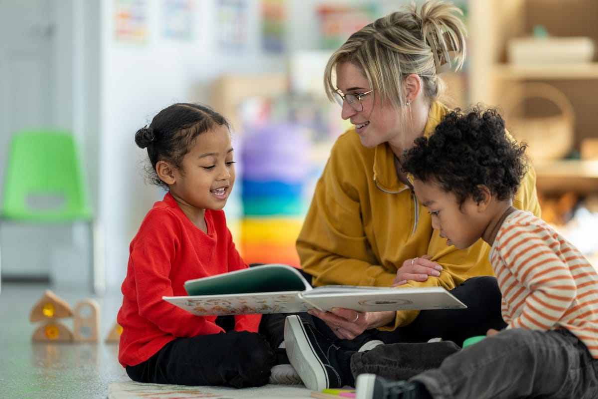 A daycare teacher sits on the floor of her classroom with a small child on either side of her, as she reads them a story.  The children are dressed comfortably and are engaged in the story.