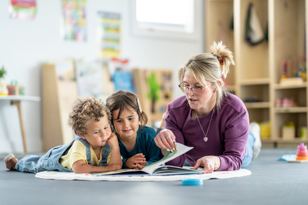 A teacher engaging with two children by reading together inside a well-lit playroom environment.