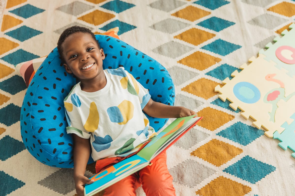 An adorable preschool age little boy smiles as he laying down in his preschool classroom and reading book.