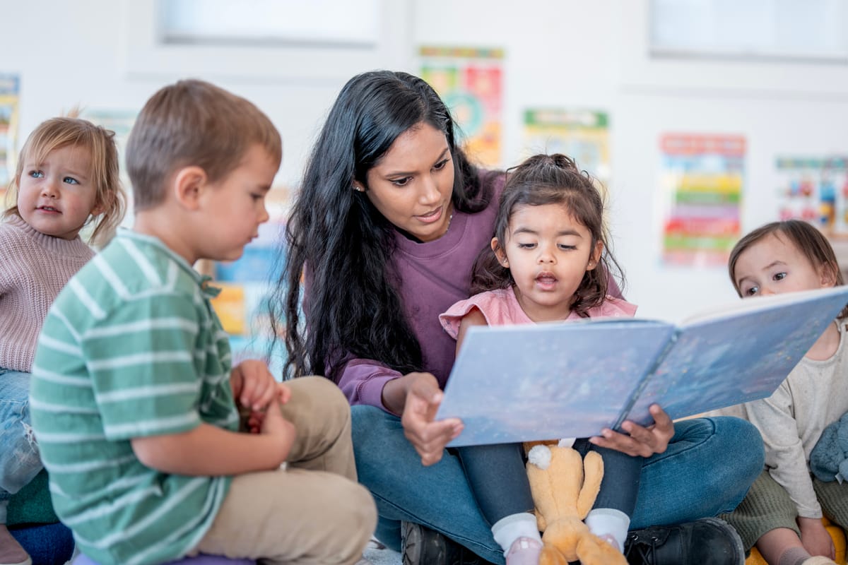 An Asian mother sits with a diverse group of smiling children as she reads a picture book in a bright classroom, fostering literacy, curiosity, and warm, family-friendly learning.