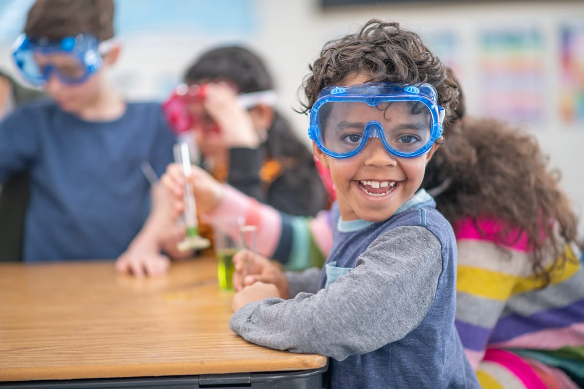 Children in a classroom participate in a science activity, wearing safety goggles and smiling. Teamwork and curiosity are emphasized as they explore science concepts in an interactive and engaging environment.