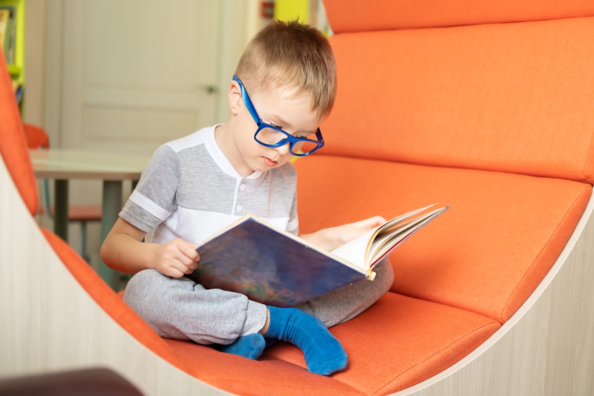 Caucasian little boy with eyeglasses reads book in modern public library. Concept of preschool and additional education, early development